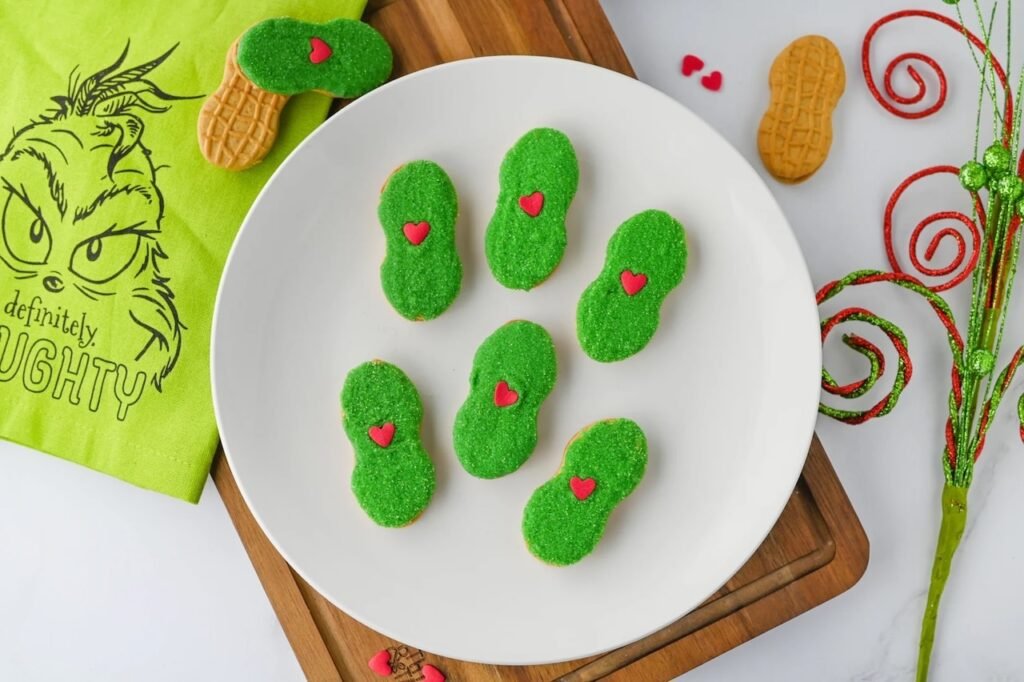 Festive Grinch Nutter Butter cookies with green candy coating and red hearts arranged on a white plate with holiday decorations.