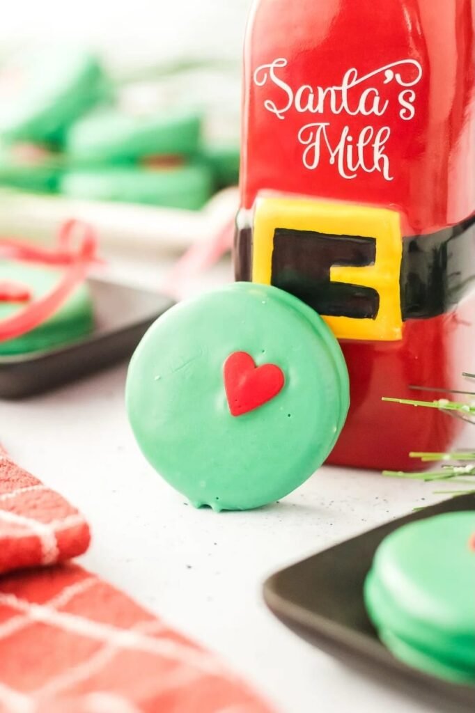 Green Grinch Oreo cookie with red heart leaning against Santa’s milk bottle, surrounded by festive holiday decor.