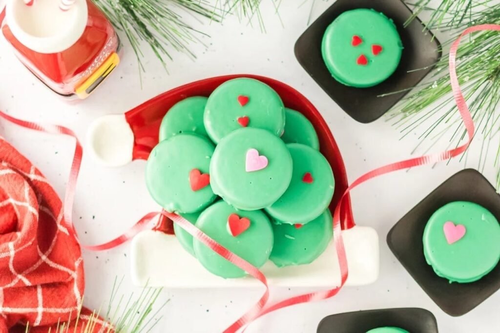 Overhead view of Grinch Oreos with red and pink hearts on a Santa hat-shaped tray surrounded by ribbon, milk bottle, and holiday greenery.