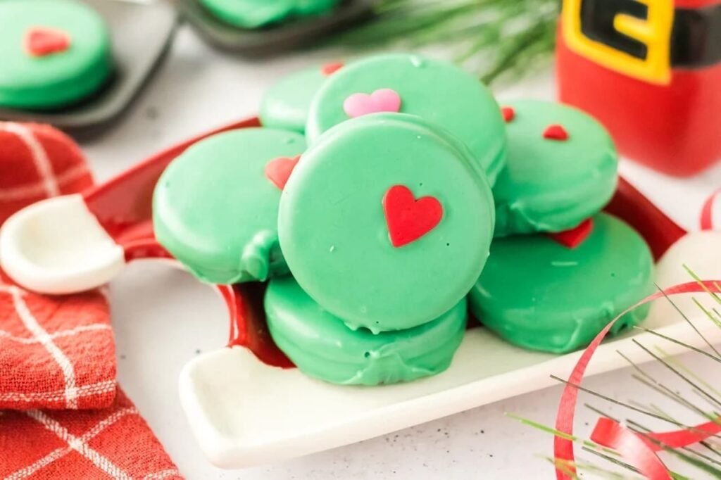stack of Grinch Oreos with red and pink heart sprinkles on a festive tray beside Santa’s milk bottle and Christmas greenery.