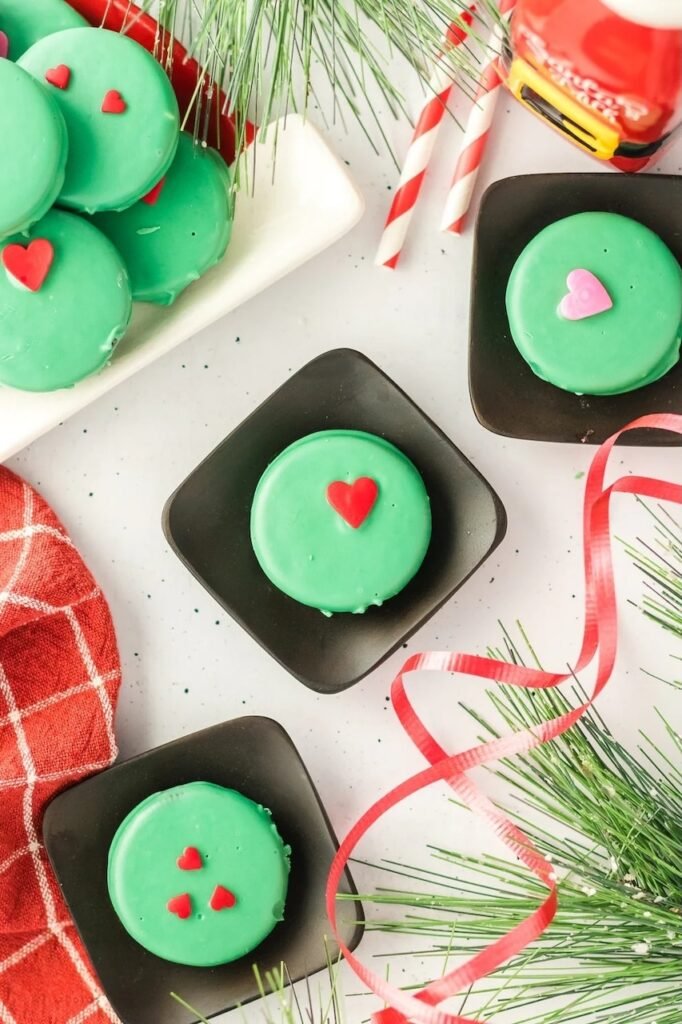 Overhead view of Grinch Oreos topped with red and pink hearts arranged on black plates with Christmas ribbon and greenery.
