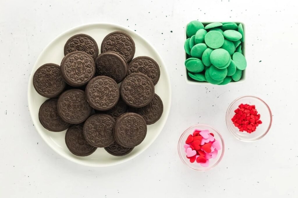 ingredients for Grinch Oreos including Oreo cookies, green candy melts, and red heart sprinkles on a white surface.