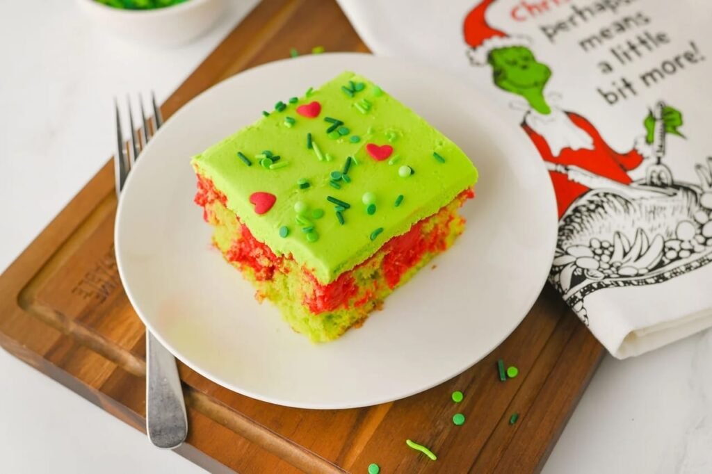 Slice of Grinch poke cake on a white plate showing bright green frosting, red pudding filling, and festive sprinkles, a fun kid-friendly Christmas dessert.