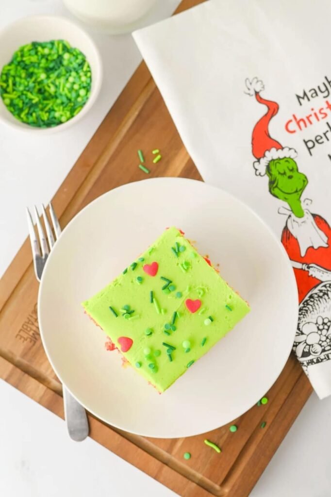 Overhead view of a slice of Grinch poke cake on a white plate with green frosting, red heart sprinkles, and a Grinch-themed napkin, a festive holiday dessert.