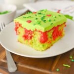 Close-up of a slice of Grinch poke cake on a white plate showing bright red pudding filling, fluffy green frosting, and festive sprinkles, an easy kid-friendly Christmas dessert.