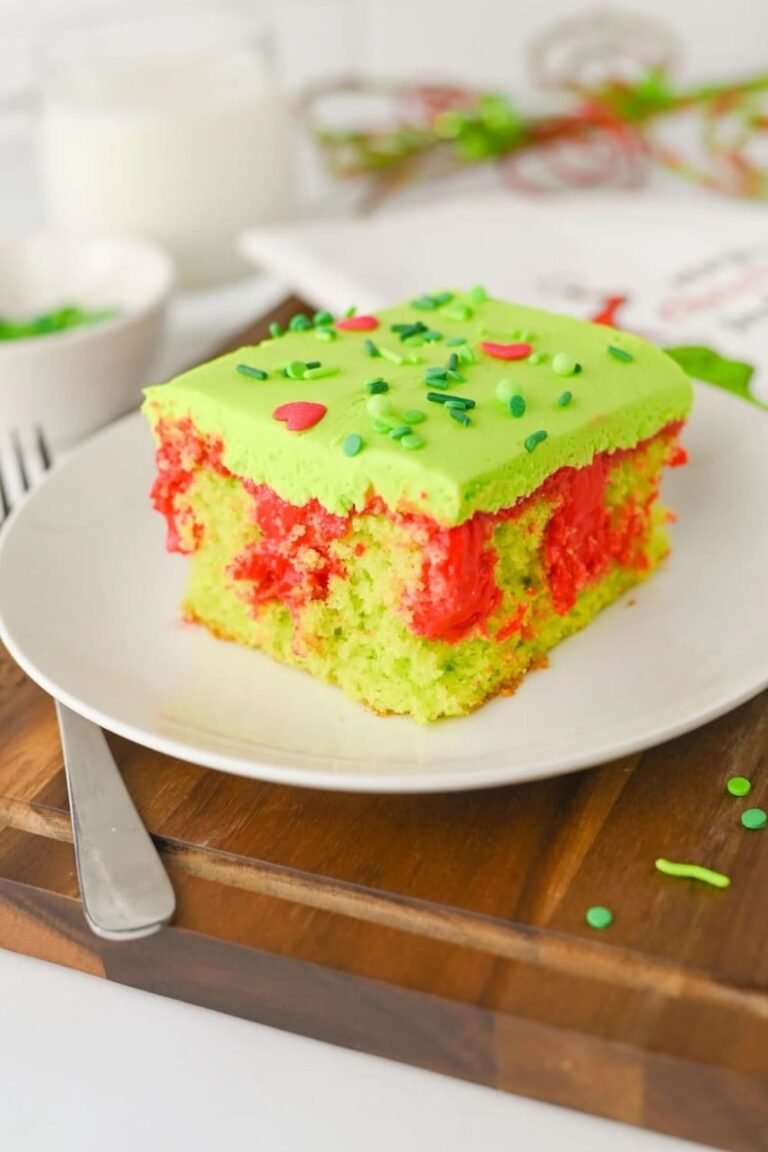 Close-up of a slice of Grinch poke cake on a white plate showing bright red pudding filling, fluffy green frosting, and festive sprinkles, an easy kid-friendly Christmas dessert.