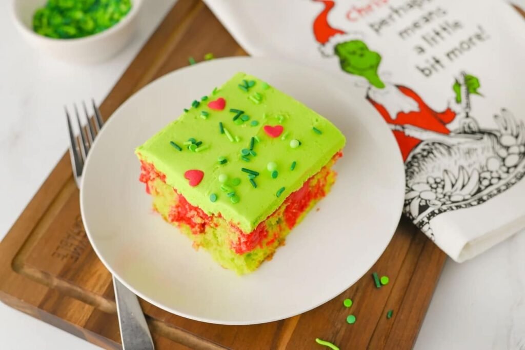 Slice of Grinch poke cake on a white plate with green frosting, red pudding filling, and festive sprinkles beside a Grinch-themed napkin, a fun holiday dessert idea.
