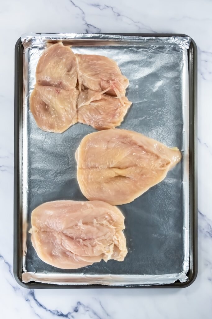 Step 1 in broiled chicken recipe – butterflied raw chicken breasts placed on a foil-lined baking sheet before seasoning.