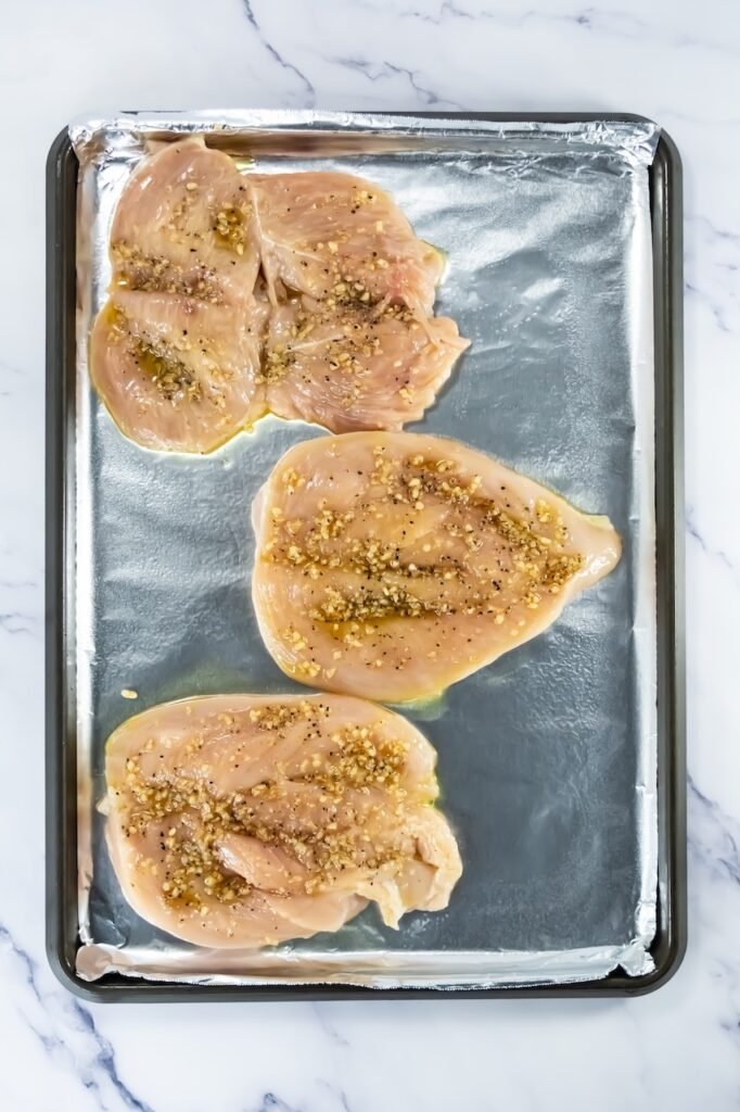 Step 4 in broiled chicken recipe – raw chicken breasts brushed with olive oil and topped with minced garlic and seasonings on a foil-lined baking sheet.