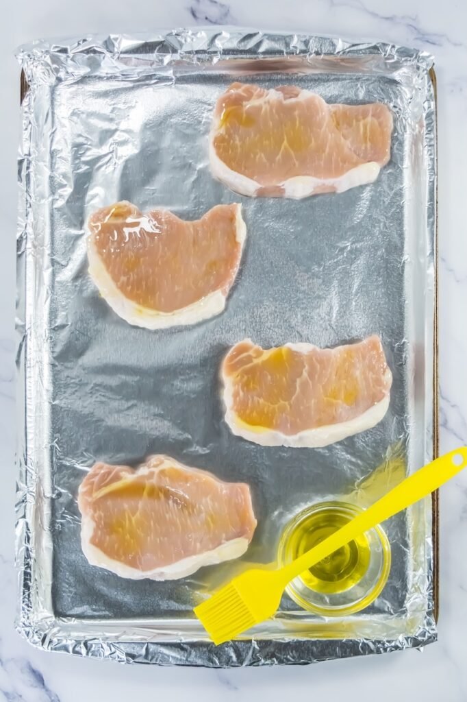 Raw pork chops on a foil-lined baking sheet brushed with olive oil using a yellow pastry brush.
