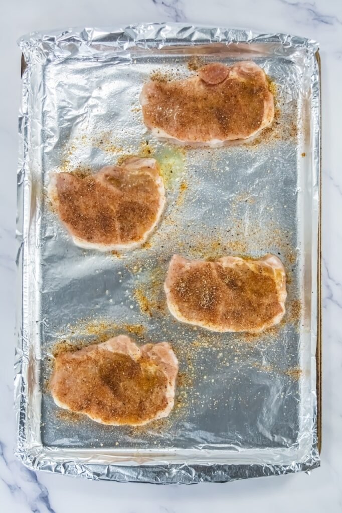 Four raw pork chops coated with seasoning on a foil-lined baking sheet, ready to broil.