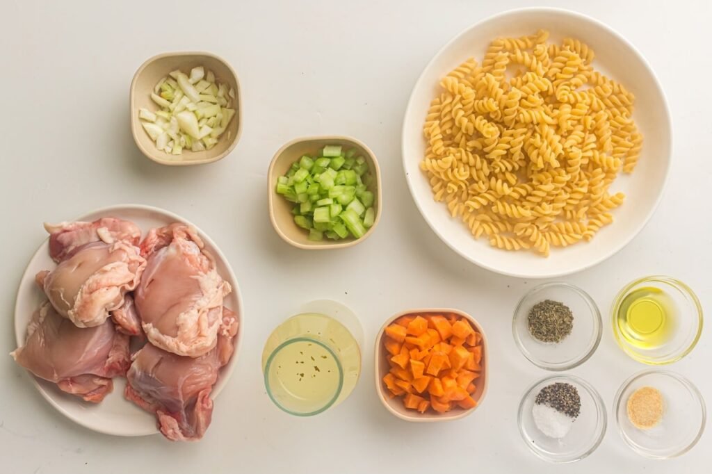 A flat lay of the ingredients for instant pot chicken noodle soup, including raw chicken thighs, fusilli pasta, chopped carrots, celery, onion, broth, olive oil, and seasonings arranged on a white surface.