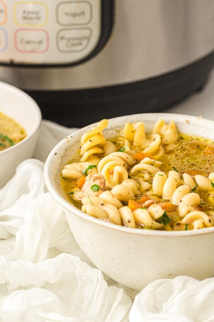 A bowl of instant pot chicken noodle soup filled with tender noodles, carrots, herbs, and shredded chicken, with an Instant Pot in the background.