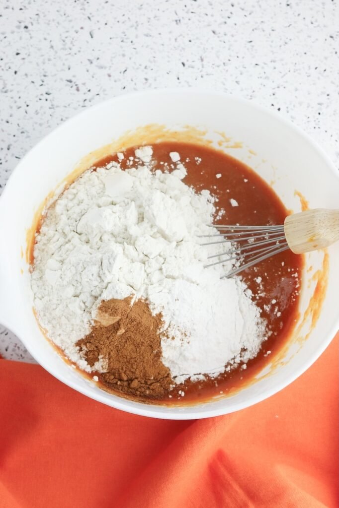 A white mixing bowl filled with pumpkin batter as flour, cinnamon, and dry ingredients are added on top, with a whisk resting in the bowl before everything is mixed together.