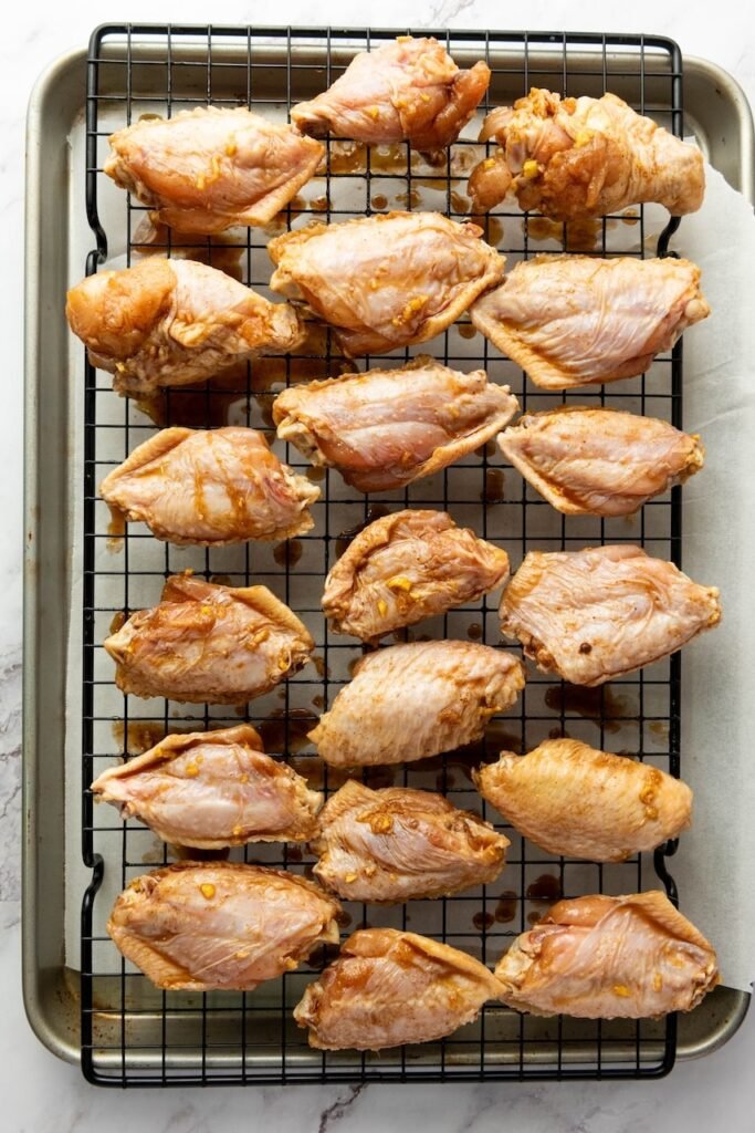Overhead photo of marinated chicken wings arranged in a single layer on a wire rack set over a foil-lined sheet pan, with small bits of garlic and ginger clinging to the wings and marinade drips on the pan below.