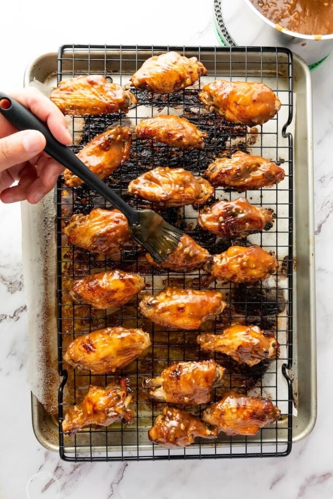 Overhead photo of baked chicken wings on a wire rack over a sheet pan while a hand uses a silicone brush to coat each wing with a thick, glossy sticky sauce; a bowl of extra glaze sits in the corner.