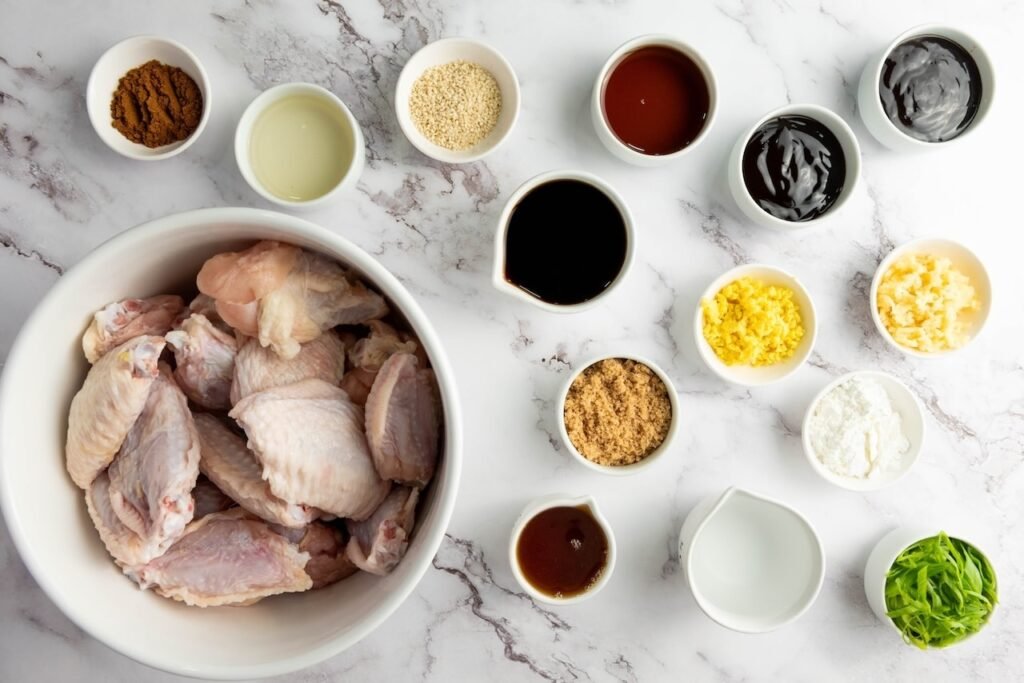 Overhead photo of raw chicken wings in a large white bowl surrounded by small bowls of marinade and sauce ingredients, including soy sauce, honey, brown sugar, vinegar, sesame seeds, minced garlic, grated ginger, cornstarch, and sliced scallions on a light marble countertop.