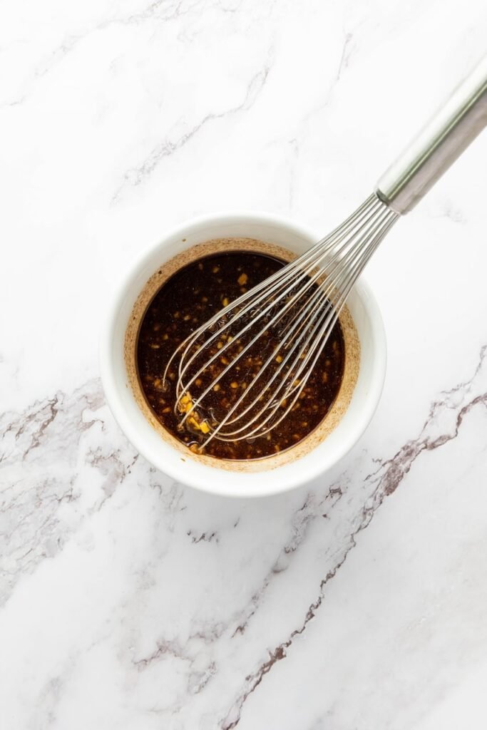 Overhead photo of a small white bowl filled with dark soy-based marinade with visible bits of minced garlic and ginger, with a metal whisk resting in the bowl on a white marble countertop.
