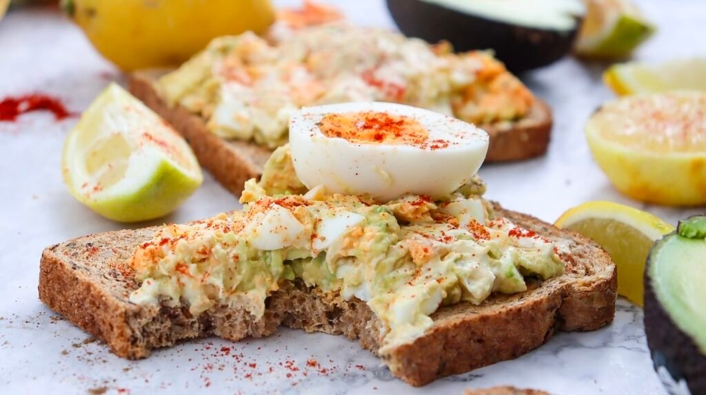 A close-up view of whole-grain toast topped with creamy avocado egg salad, finished with a sliced hard-boiled egg and light seasoning, with lemon wedges and avocado in the background.