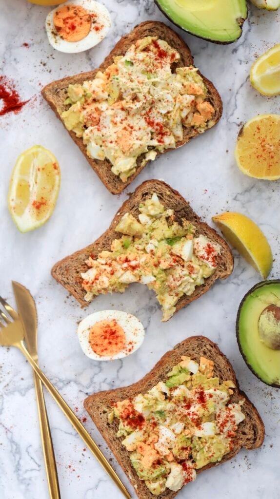 Slices of whole-grain toast are topped with creamy avocado egg salad and sprinkled with seasoning, arranged on a light surface with lemon wedges, avocado halves, and sliced hard-boiled eggs around them.