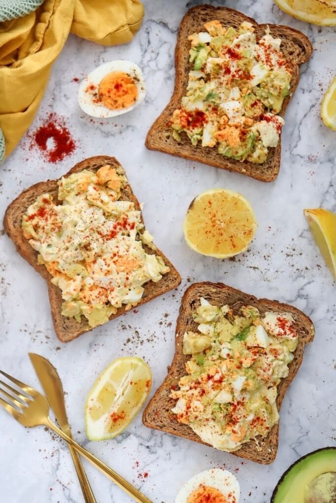 Several slices of whole-grain toast are topped with creamy avocado egg salad and lightly sprinkled with seasoning, arranged on a marble surface with lemon wedges, avocado halves, and sliced hard-boiled eggs nearby.