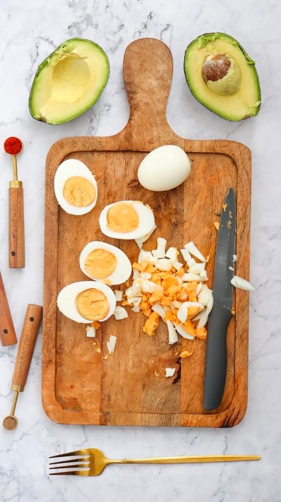 Hard-boiled eggs chopped on a wooden cutting board alongside halved avocados, a knife, and small measuring spoons, showing the fresh ingredients used to make avocado egg salad before mixing.