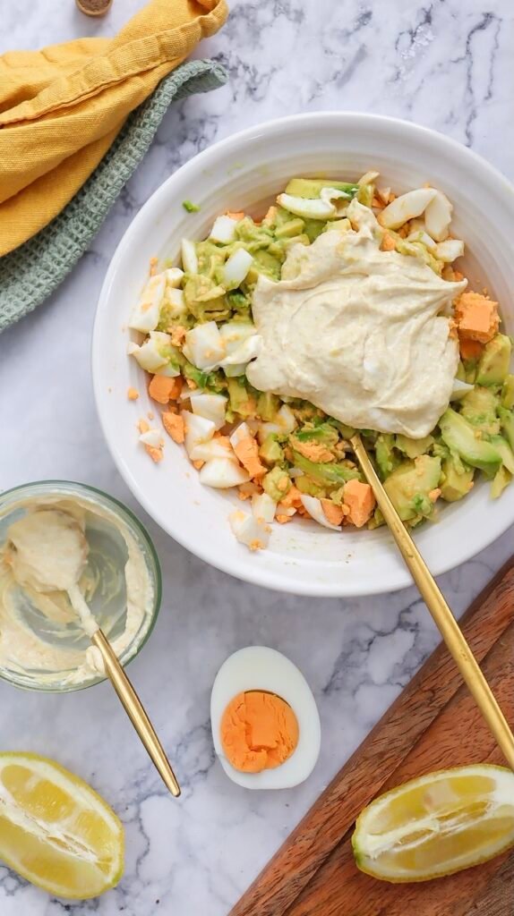 Chopped hard-boiled eggs and mashed avocado are mixed in a white bowl with a spoonful of Greek yogurt added on top, before it is fully combined.