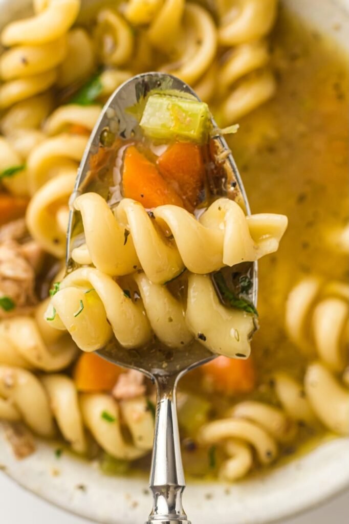 Close-up of a spoonful of chicken noodle soup with spiral noodles, diced carrots, celery, and herbs lifted from the bowl.