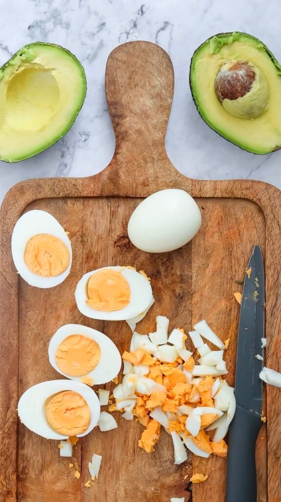 Hard-boiled eggs are sliced and chopped on a wooden cutting board with a knife, while halved avocados sit above, showing the egg prep step before making.