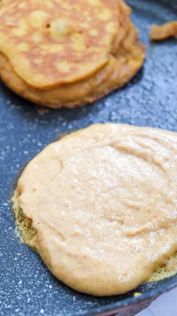 Close-up view of pumpkin pancake batter cooking on a non-stick griddle, showing bubbles forming on the surface as the pancake cooks and begins to set before flipping.