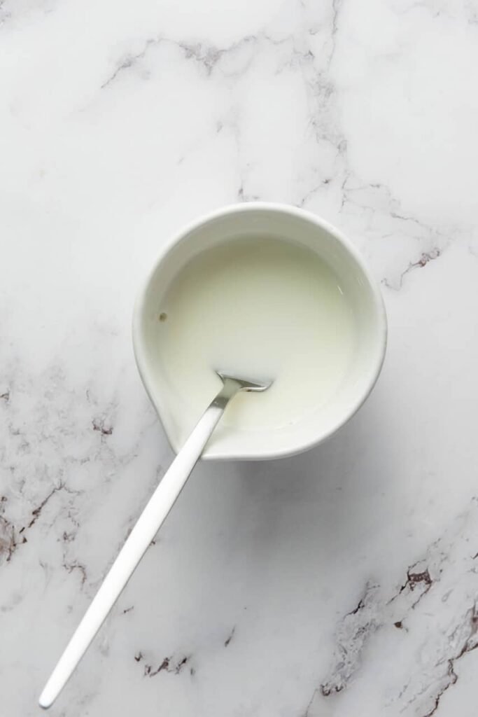 Overhead photo of a small white bowl filled with a pale, milky cornstarch-and-water slurry, with a spoon resting inside the bowl on a light marble countertop.