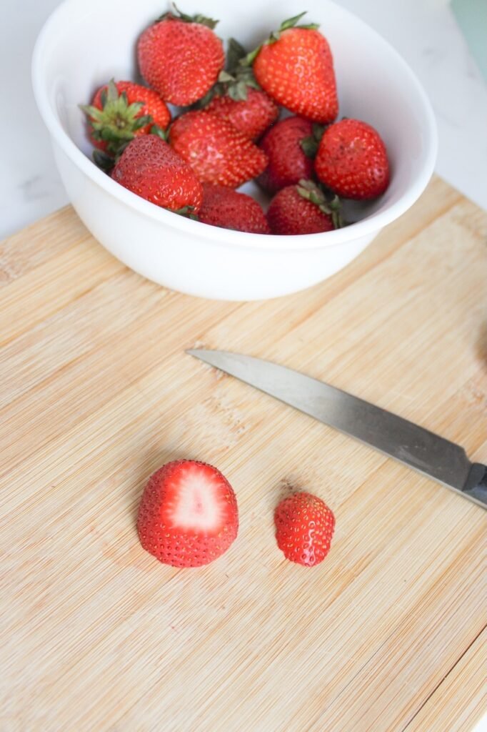 A cutting board with a knife, a bowl of whole strawberries, and one strawberry sliced into a flat-bottomed base and a small top piece, showing how to prepare the fruit for assembling.
