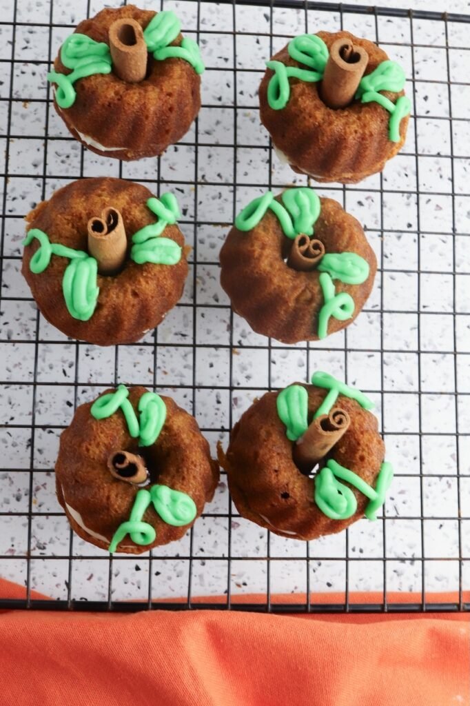 Finished desserts decorated with green frosting vines and cinnamon stick stems, arranged on a wire cooling rack and styled to resemble small pumpkins.