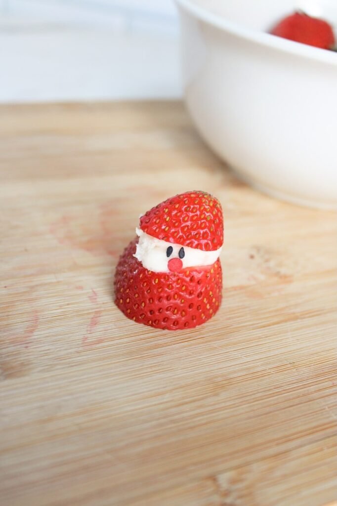 A partially assembled Christmas dessert on a wooden cutting board, filled with white chocolate cream cheese and decorated with two small black sprinkle eyes and a red sprinkle nose, showing the step where the face is added.