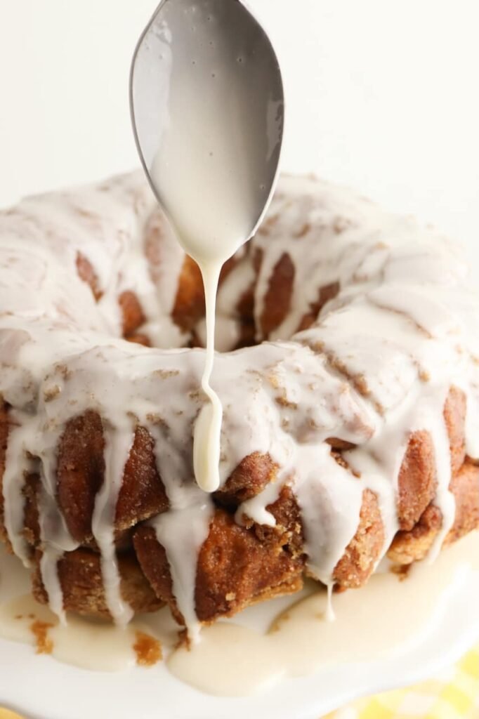Vanilla glaze being drizzled from a spoon over a baked dessert on a white cake stand