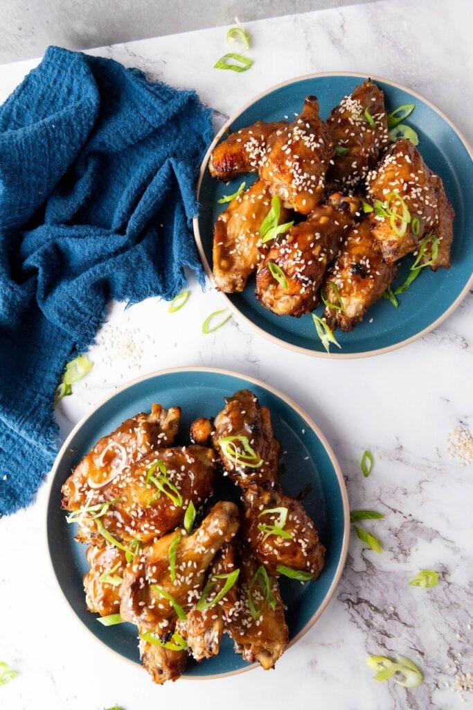 Overhead photo of two blue plates filled with Asian sticky chicken wings coated in a shiny glaze and topped with sesame seeds and sliced green onions, with a dark blue cloth napkin and scattered garnishes on a light countertop.