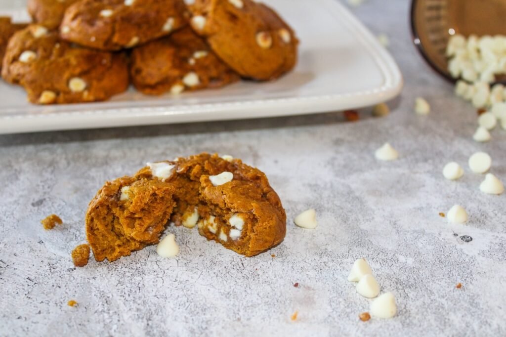 A broken pumpkin spice cookie sits on a light gray countertop, showing a soft, cake-like interior with white cream cheese-flavored baking chips melted inside. More baking chips and cookie crumbs are scattered nearby, and a tray of additional cookies is blurred in the background, highlighting the cookies’ tender texture.