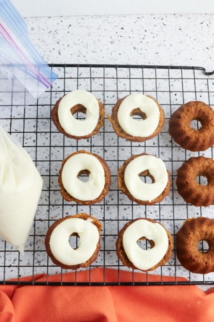 Pumpkin mini Bundt cakes resting on a wire cooling rack as cream cheese frosting is piped onto the flat side of each cake before being assembled into sandwich-style cakes.