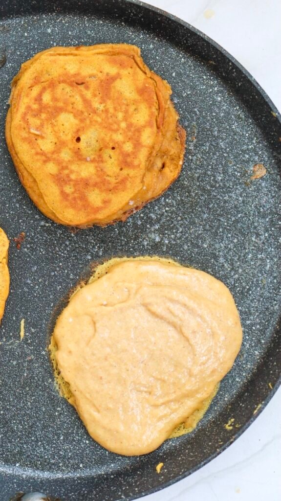 Overhead view of two pumpkin pancakes cooking in a non-stick skillet, with one pancake flipped and golden brown while the other continues cooking on the first side, showing the proper stage for flipping.