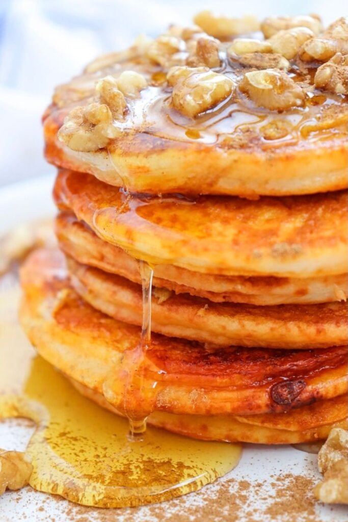 Close-up view of a tall stack of fluffy pumpkin pancakes topped with chopped walnuts and warm maple syrup dripping down the sides, served on a white plate with cinnamon sprinkled underneath for a cozy fall breakfast.