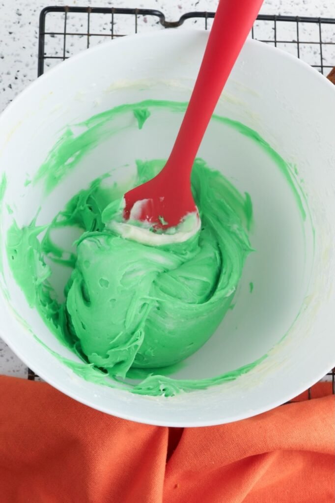 A white mixing bowl with cream cheese frosting tinted green using food coloring, being stirred with a spatula to prepare frosting for decorating.