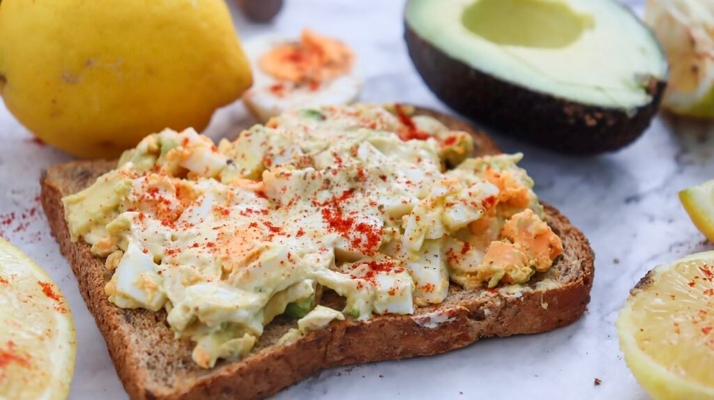 A close-up view of whole-grain toast topped with creamy avocado egg salad and a dusting of seasoning, with lemon wedges and avocado halves visible in the background.