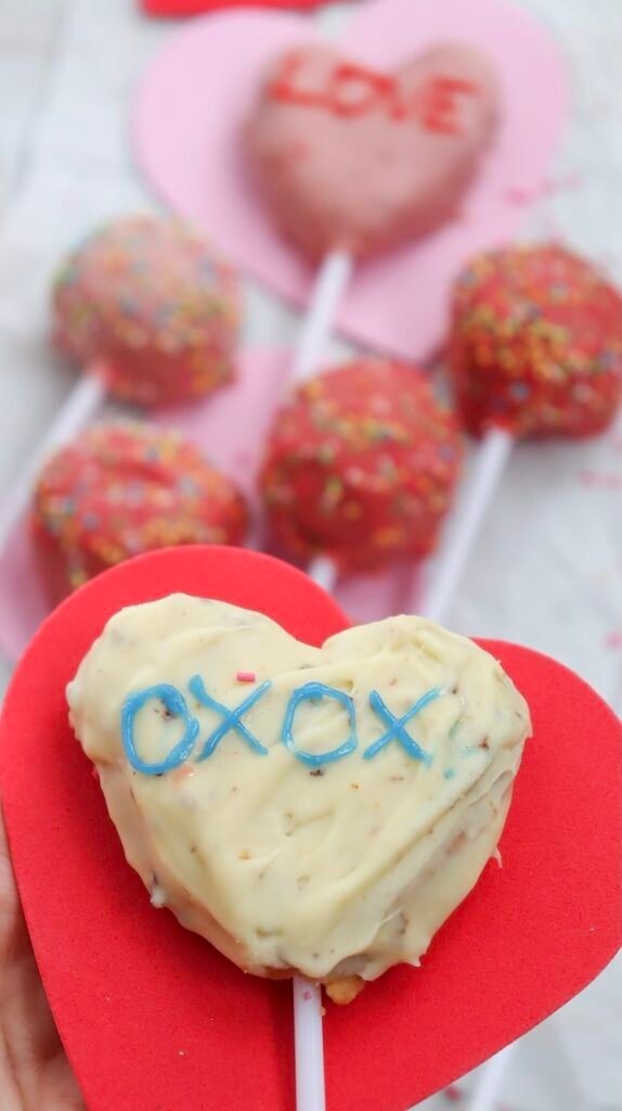 Close-up photo of a heart-shaped white chocolate Valentine pop with an XOXO message, shown with colorful sprinkle pops in the background.