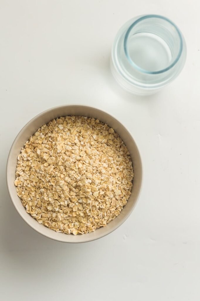 A bowl filled with old-fashioned rolled oats sitting on a white countertop next to a small glass jar of water, showing the basic ingredients needed to make creamy Instant Pot oatmeal before cooking.