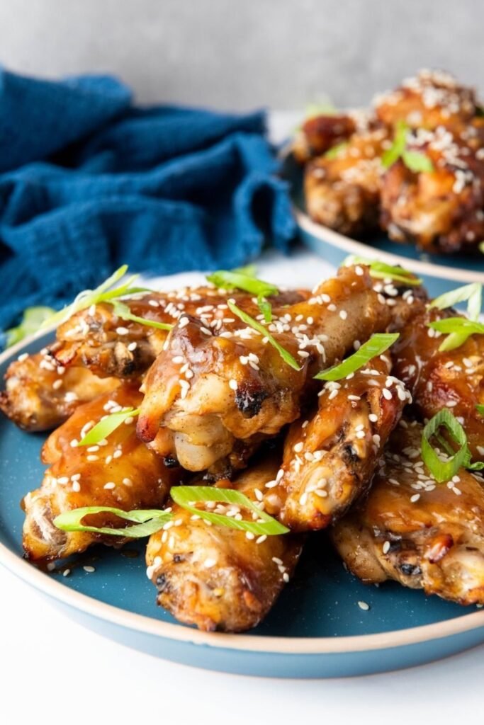 Close-up of Asian sticky chicken wings coated in a shiny honey-soy glaze on a blue plate, topped with toasted sesame seeds and sliced green onions, with a second plate of wings blurred in the background.