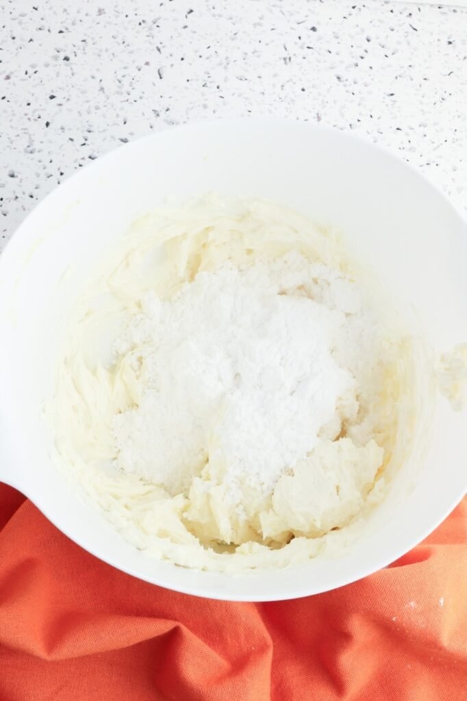 A white mixing bowl with softened cream cheese and butter being mixed with powdered sugar to make a smooth cream cheese frosting.