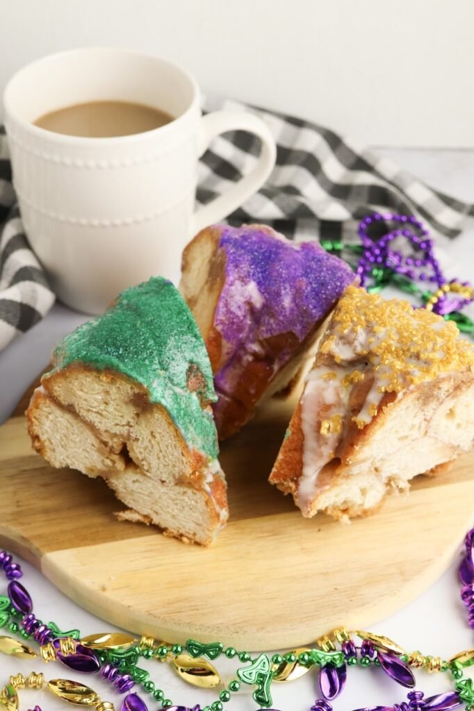 Three colorful slices of Mardi Gras King Cake with purple, green, and gold sugar served on a wooden board next to a mug of coffee and festive beads