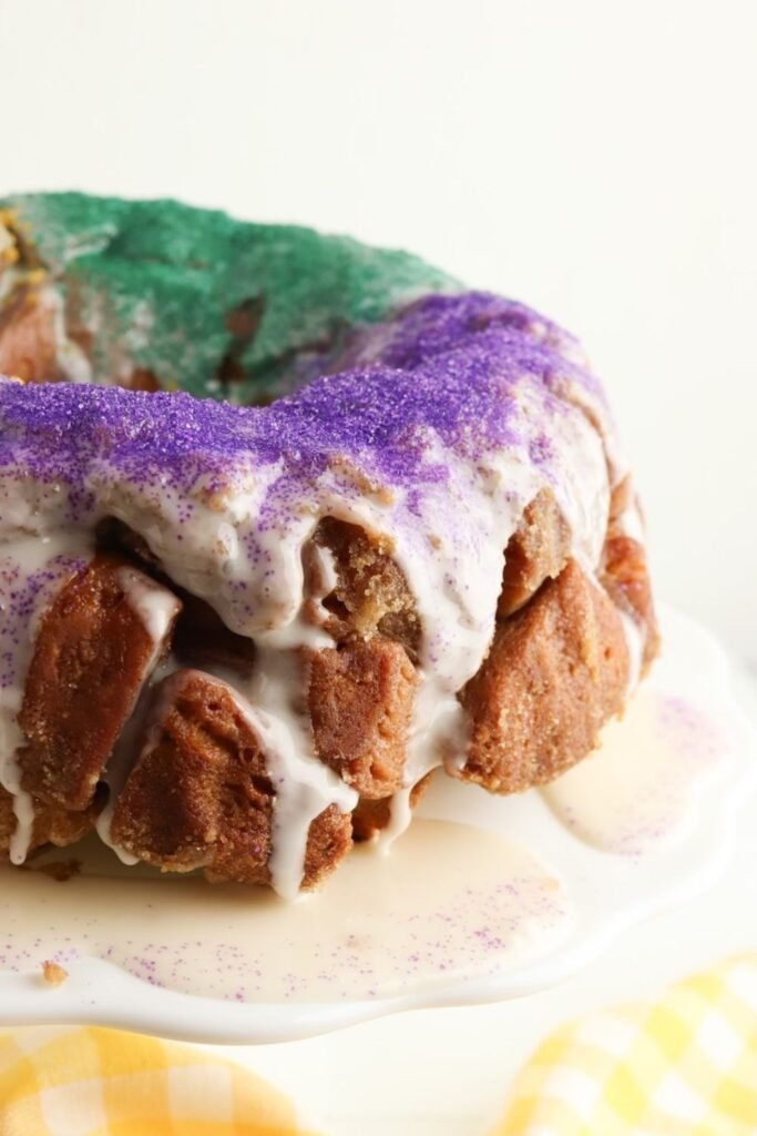 Pull apart King Cake with purple and green sugar sprinkles, iced and ready to serve on a white cake stand.