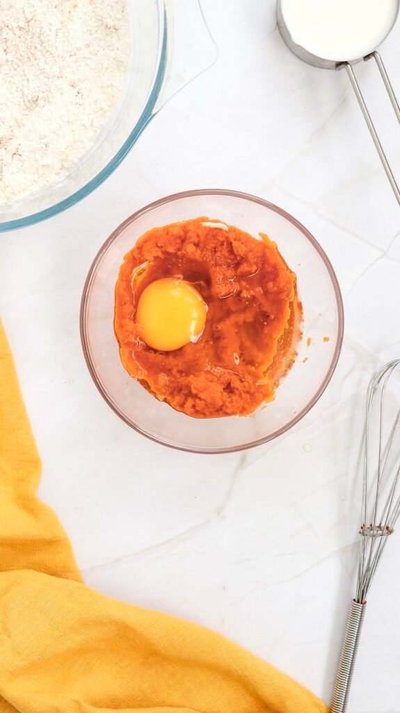 Overhead view of a bowl with pumpkin purée, a cracked egg, and liquid ingredients being mixed together, showing the wet ingredients prepared before combining with the dry ingredients for pumpkin pancake batter.