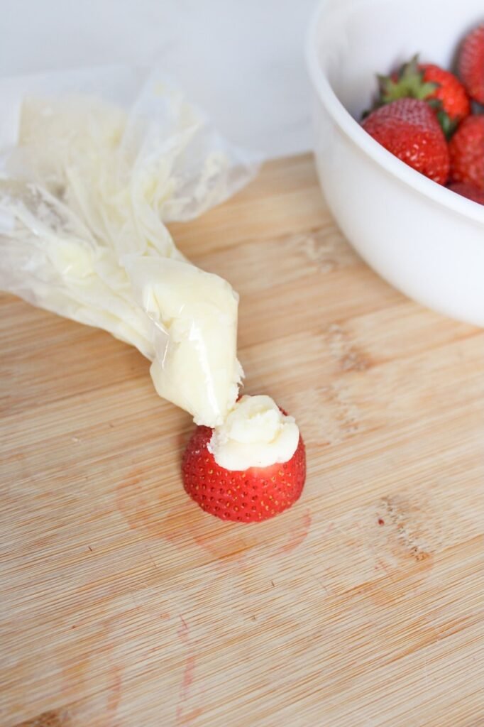 A piping bag filled with a white chocolate and cream cheese mixture is shown piping a swirl of filling onto the bottom half of a sliced strawberry on a wooden cutting board, with a bowl of whole strawberries beside it.